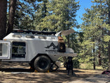 Mammoth Overland RV rental: A rugged trailer with a happy black dog awaits adventure in a sunny pine forest campsite.