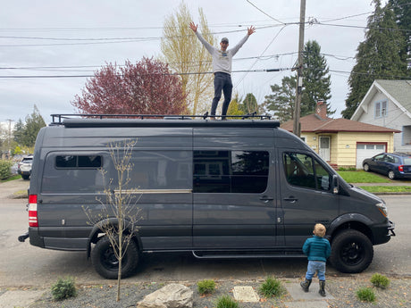 Man joyfully stands atop a sleek grey camper van (RV rental) with a child admiring it. Perfect for your next adventure!