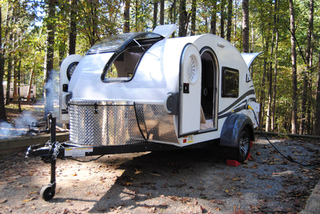 This modern teardrop RV, perfect for an **RV rental**, is set up at a wooded campsite with smoke from a fire and another camper.