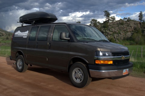 Dark grey Basecamper van RV rental with roof box, parked on a dirt road. Rocky hills and cloudy sky in background. — photo 2