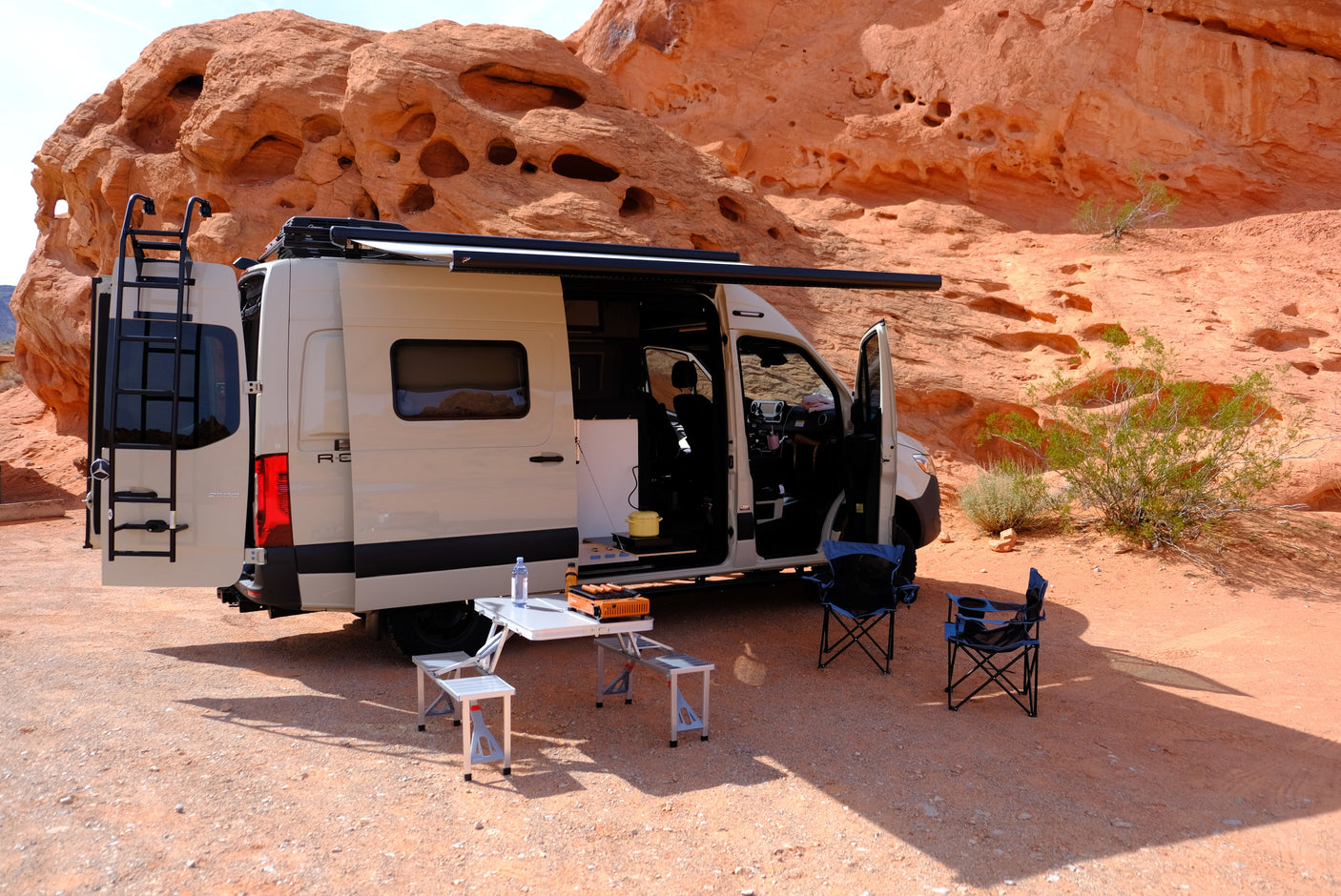 RV rental camper van parked in a desert landscape with red rock formations, set up with awning, table, & chairs.