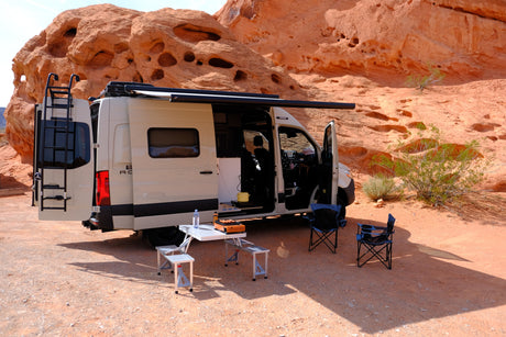 RV rental camper van parked in a desert landscape with red rock formations, set up with awning, table, & chairs.