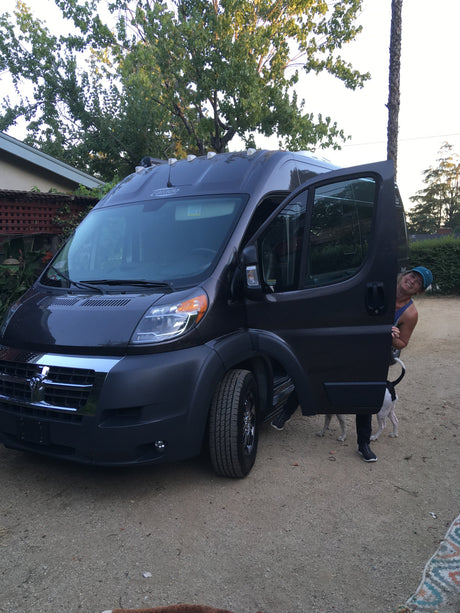 Smiling person and dog peeking from an open side door of a dark grey RV rental, parked on a dirt lot.