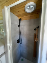 Modern tiled shower in a compact RV rental, featuring a black shower head, wood ceiling, and hanging bath accessories.