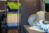 Inside an RV rental: a plush lounge, elevated bunk, and the rear open to a dirt road & green hills.