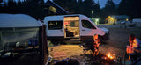A cozy evening with an RV rental camper van, pop-top tent open, and friends by the campfire at a busy campground. Classic van life.