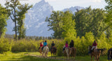 A group of horseback riders traverses a green meadow with majestic, snow-capped mountains. An RV rental enhances outdoor adventures.