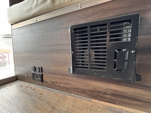 An RV rental interior shows a black furnace vent, USB ports, and electrical outlets on a wood-grain panel.