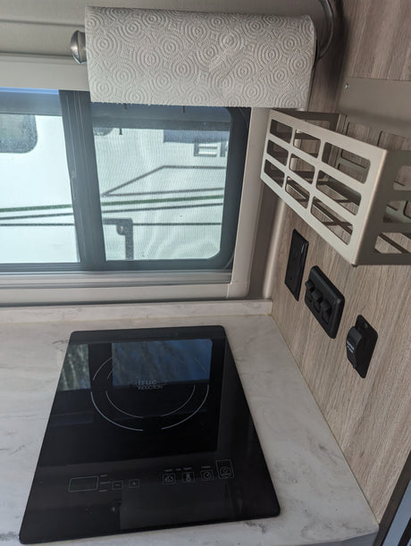 Modern rv rental interior: induction cooktop, paper towels, window view, and water pump/light controls on wood panel.