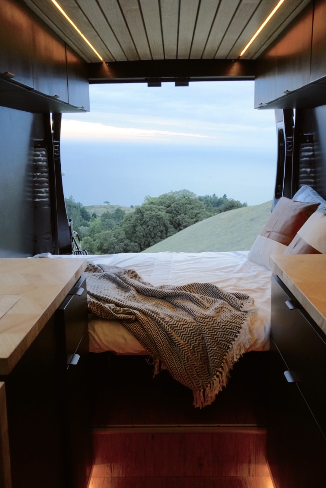 Cozy rv rental campervan interior with bed, blanket, and wood trim. Scenic view of green hills and ocean from the rear doors. — photo 3