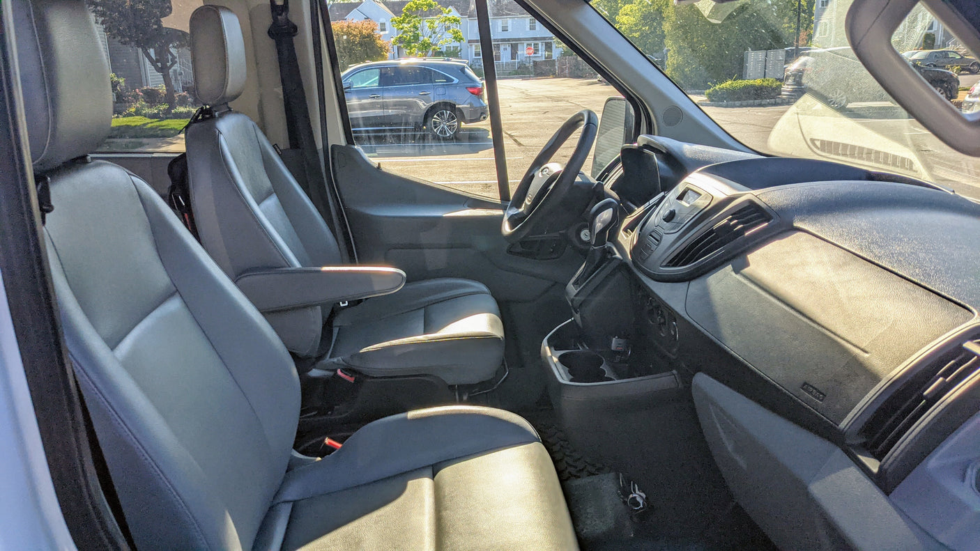 Interior view of an RV rental van, showing the gray front seats, dashboard, and steering wheel, with a sunny street scene outside.
