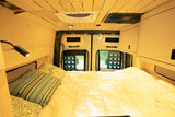 Inside a cozy RV rental: a bed with soft bedding, warm wood paneling, overhead storage, and ceiling climate units.