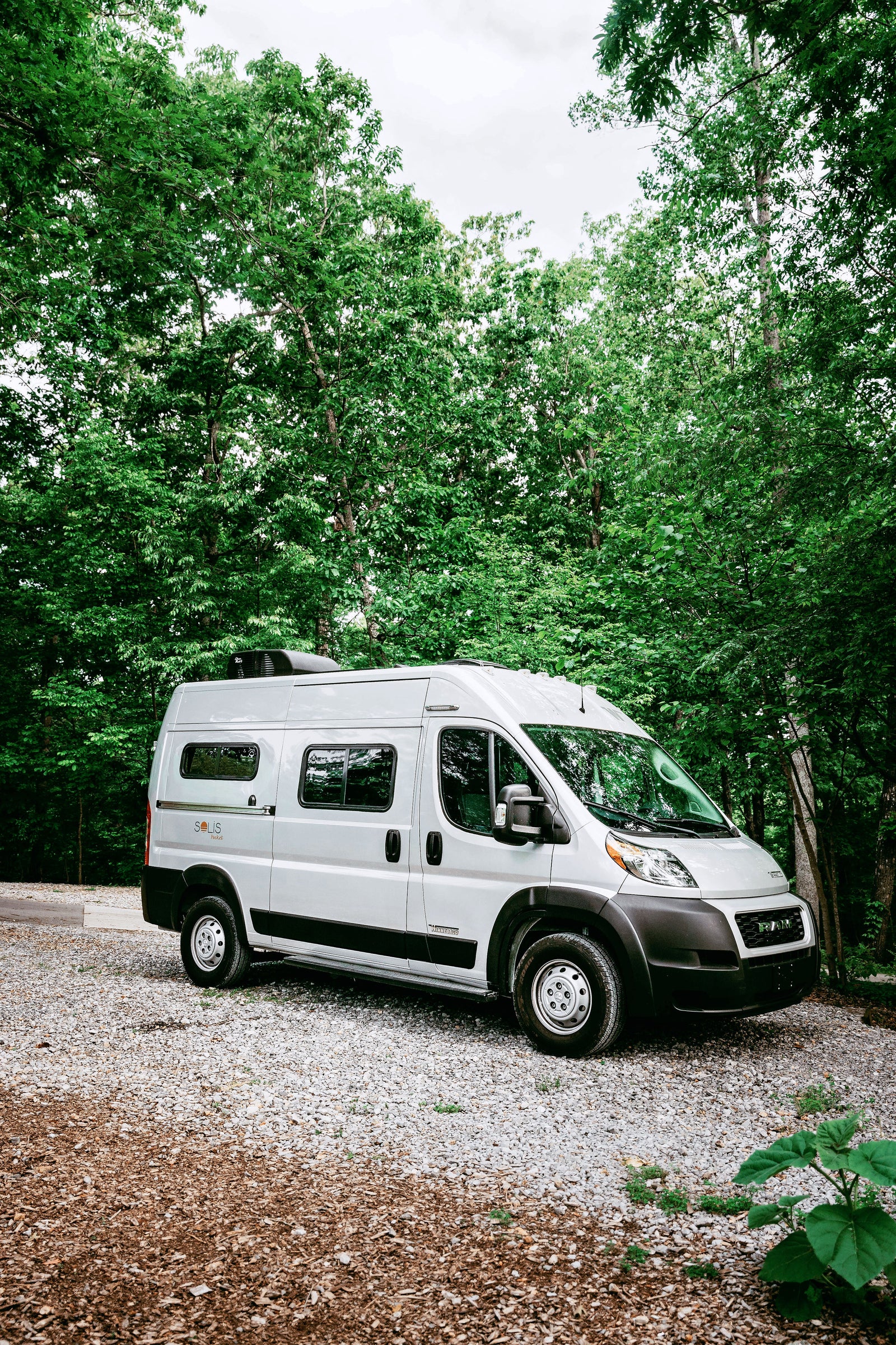 A sleek Winnebago Solis RV rental, silver, parked on a gravel lot surrounded by lush green trees, ready for adventure.