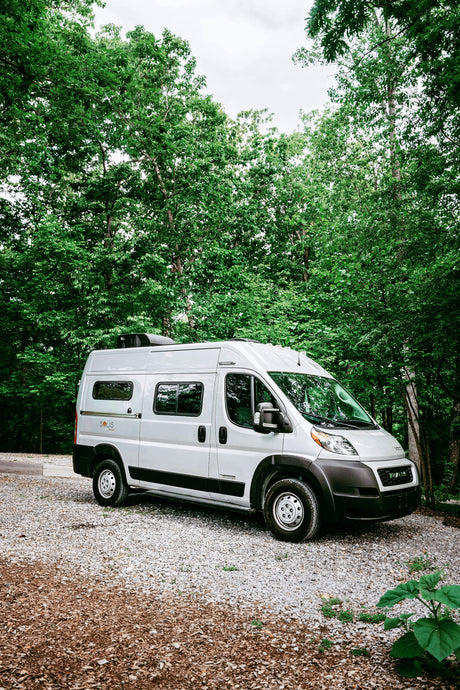 A sleek Winnebago Solis RV rental, silver, parked on a gravel lot surrounded by lush green trees, ready for adventure.