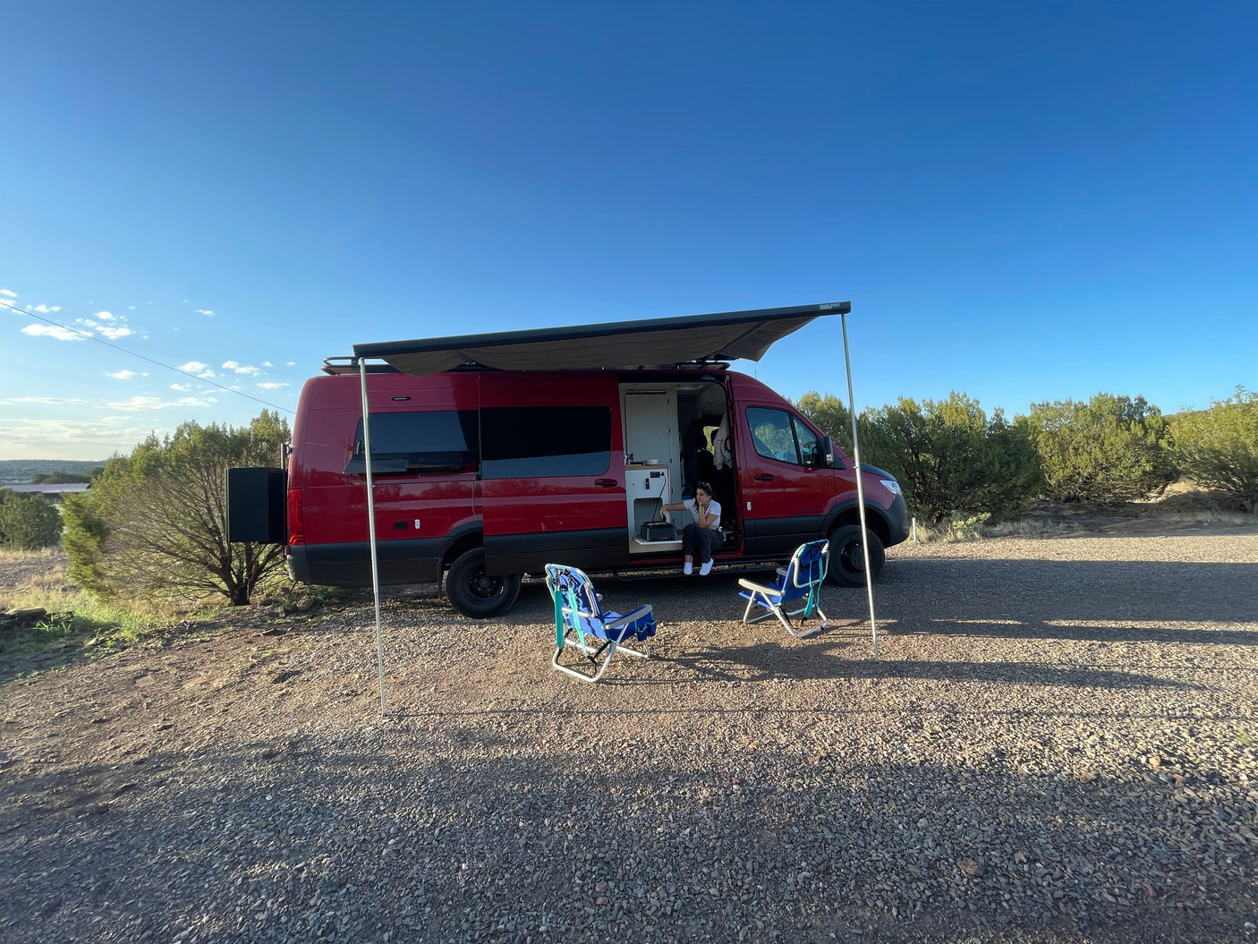 Relaxing outdoors in a red RV rental van with its awning extended, a person sits inside, and chairs await on the gravel.