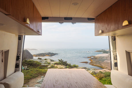 Looking out from an RV rental: a serene coastal view with rocky shores. Inside, wooden cabinets & table are visible.
