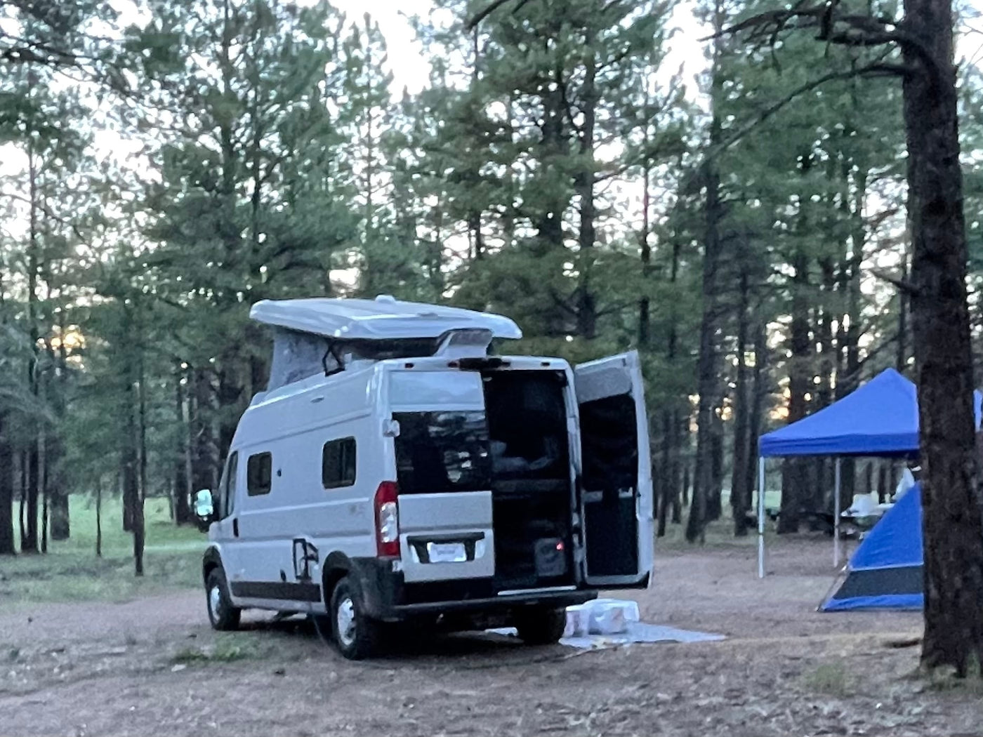 An RV rental camper van with open back doors is set up for camping in a forest site, near a blue tent and canopy. — photo 7
