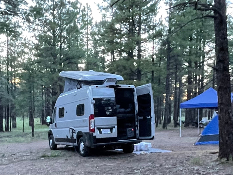 An RV rental camper van with open back doors is set up for camping in a forest site, near a blue tent and canopy. — photo 7