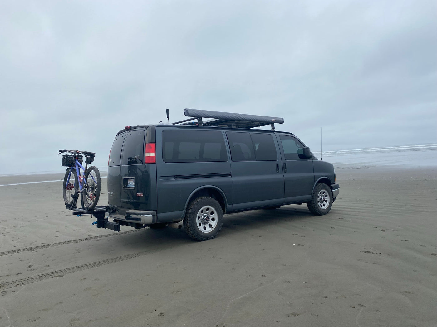 A gray GMC rv rental van with a bike rack and roof awning is parked on a vast sandy beach beside the ocean. — photo 19