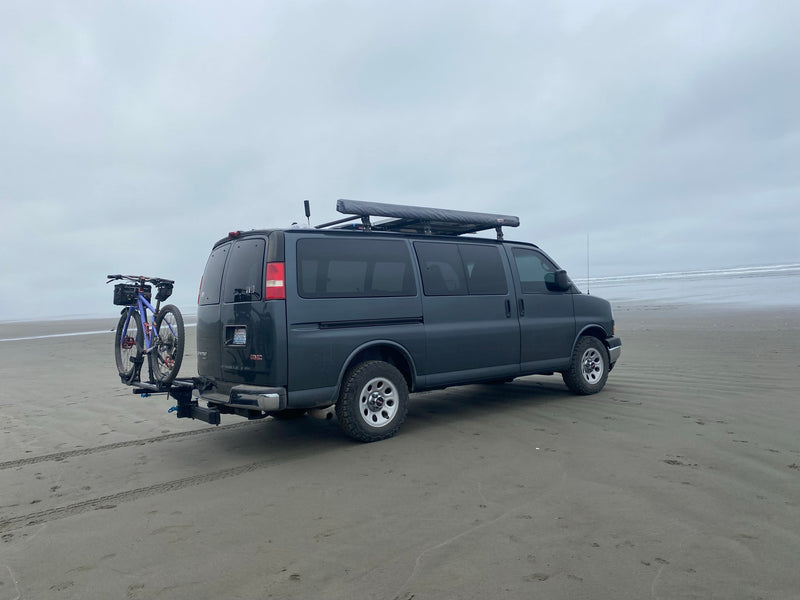 A gray GMC rv rental van with a bike rack and roof awning is parked on a vast sandy beach beside the ocean. — photo 19