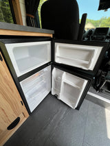 This RV rental features a clean, open compact refrigerator with empty freezer and fridge sections, ready for use.