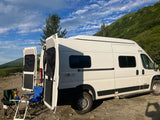 Scenic RV rental adventure! White camper van with open rear doors, outdoor table & chairs, set against green mountains.