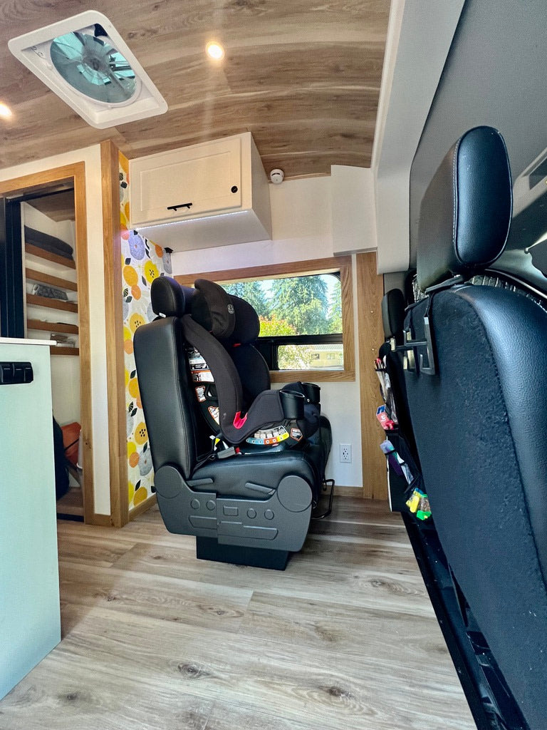 Bright rv rental interior with wood-look flooring, a child's car seat, overhead cabinets, and a ceiling vent.