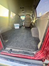 Interior of an RV rental's cargo area, showcasing a dark carpeted floor and a white RinseKit cooler. Front seats visible ahead.