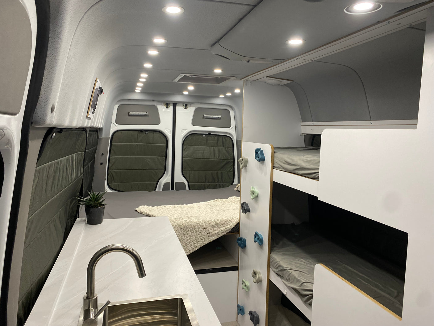 This bright RV rental interior features a kitchen sink, rear bed, and bunk beds with playful climbing holds for a unique family travel experience.