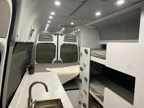 This bright RV rental interior features a kitchen sink, rear bed, and bunk beds with playful climbing holds for a unique family travel experience.