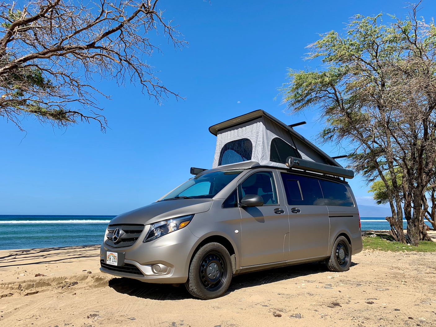 Explore Hawaii with this RV rental! A pop-top camper van on a sandy beach next to the ocean under a blue sky.