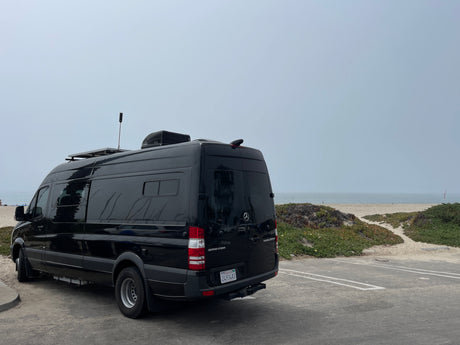 Black Sprinter rv rental parked at a serene beachside lot. Ocean, sandy path, and hazy sky visible, perfect for coastal adventures.