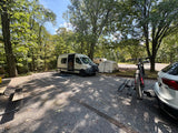 An RV rental camper van, tent, and mountain bike are set up at a shaded, wooded campsite for a relaxing trip.