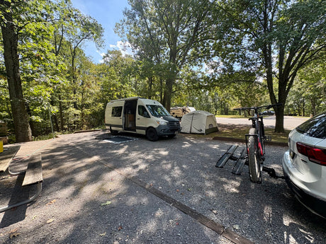 An RV rental camper van, tent, and mountain bike are set up at a shaded, wooded campsite for a relaxing trip.