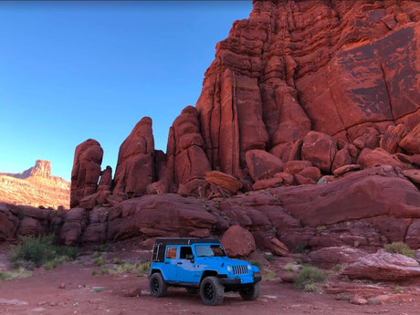 A blue Jeep, an RV rental equipped for overlanding, rests among towering red rock formations in a vast desert landscape.