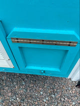 Detailed view of a teal RV rental panel with a brass hinge and latch. White louvered vent to left, gravel below.