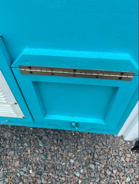 Detailed view of a teal RV rental panel with a brass hinge and latch. White louvered vent to left, gravel below.