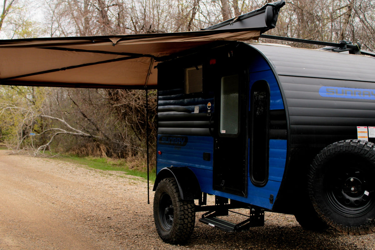 A blue and black teardrop camper RV rental with an extended awning, parked on a gravel path amidst trees. Ready for adventure. — photo 2