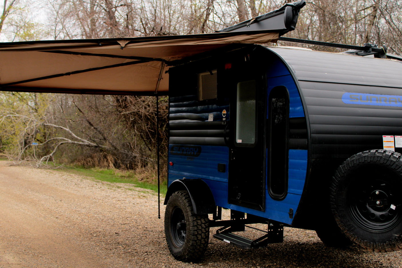 A blue and black teardrop camper RV rental with an extended awning, parked on a gravel path amidst trees. Ready for adventure.