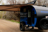 A blue and black teardrop camper RV rental with an extended awning, parked on a gravel path amidst trees. Ready for adventure.