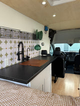 Modern camper van kitchen in an RV rental, with dark counter, patterned backsplash, black faucet, and glimpse of the driver's cabin.
