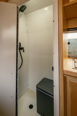 This rv rental features a compact shower with a portable toilet, overhead shower, and drain. Adjacent kitchen and scenic window view.