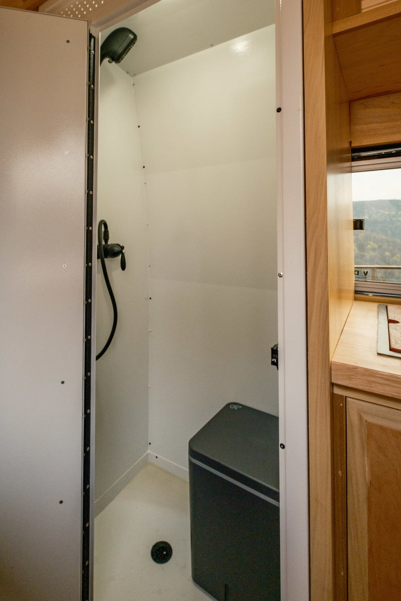 This rv rental features a compact shower with a portable toilet, overhead shower, and drain. Adjacent kitchen and scenic window view. — photo 10