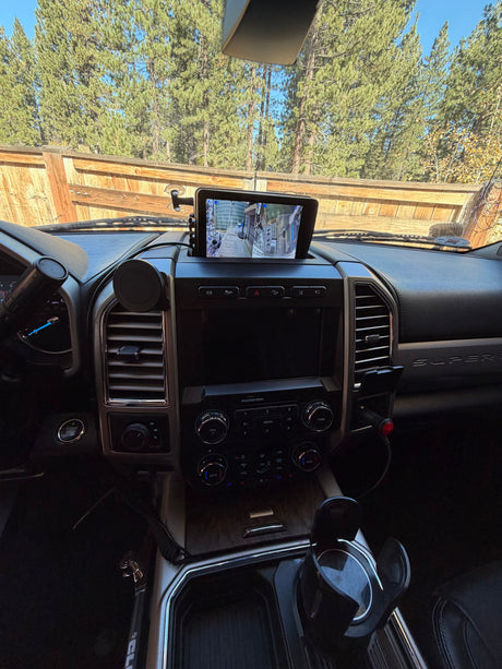 Inside a Ford Super Duty RV rental truck: dashboard, camera display showing a towed camper, with pine trees visible outside.