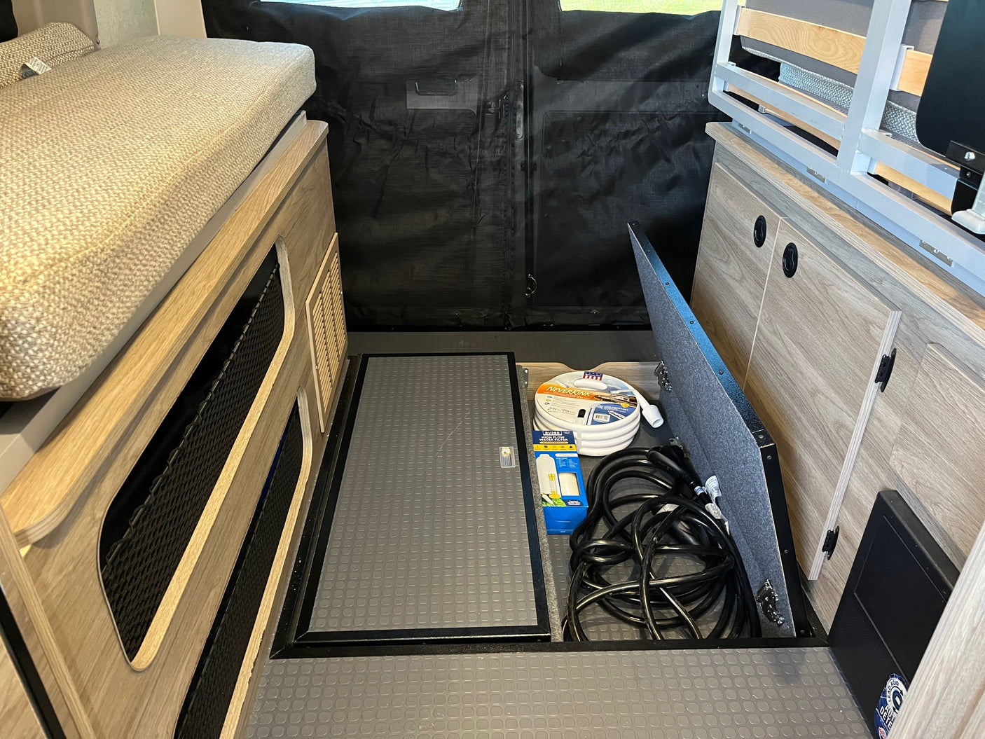 RV rental interior reveals a bed, wood cabinets, and floor storage with a white water hose, filter, and black electrical cords.