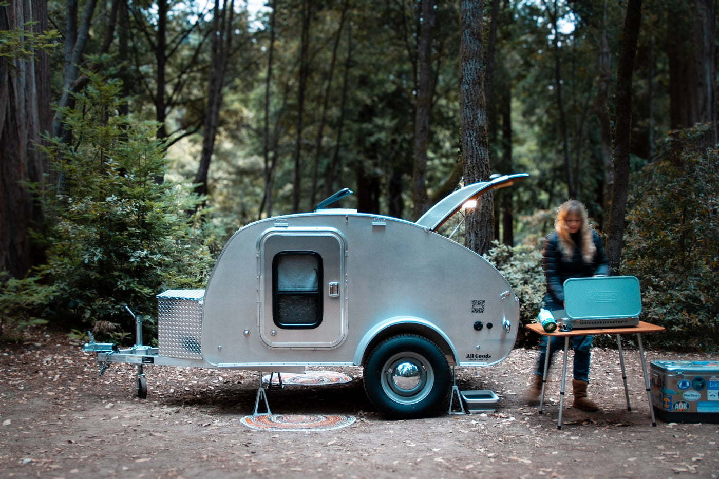 Camping setup with a sleek teardrop RV rental parked among tall trees. A person prepares a meal on a stove next to it.|Camping rv rental: A sleek teardrop trailer with an open rear kitchen — photo 8
