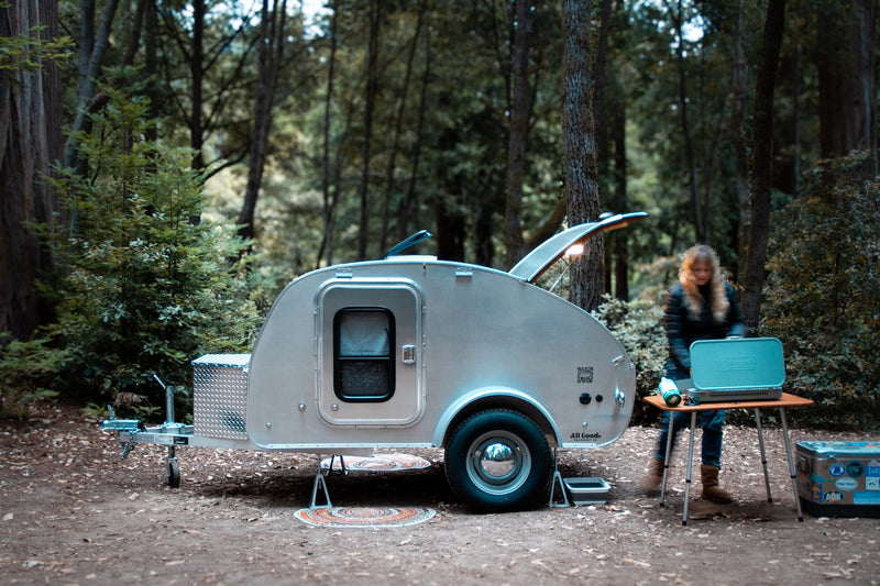 Camping setup with a sleek teardrop RV rental parked among tall trees. A person prepares a meal on a stove next to it.|Camping rv rental: A sleek teardrop trailer with an open rear kitchen — photo 8