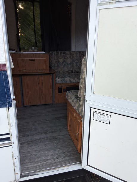 Interior of an rv rental camper, featuring a kitchenette, dinette, and wood floor. Seen from the open entry door with a gas safety warning.