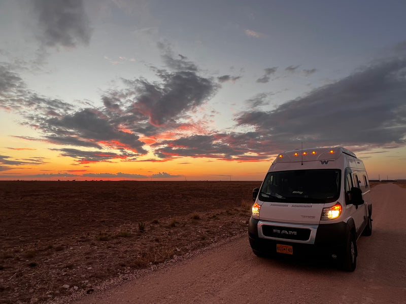 Embrace the open road! This white rv rental camper van illuminates a dirt road at stunning sunset. Field to left.|RV rental parked at dusk by the ocean — photo 1