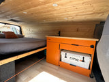 This RV rental showcases a custom interior with a bed, wood paneling, and an orange kitchen cabinet storing an RTIC cooler.